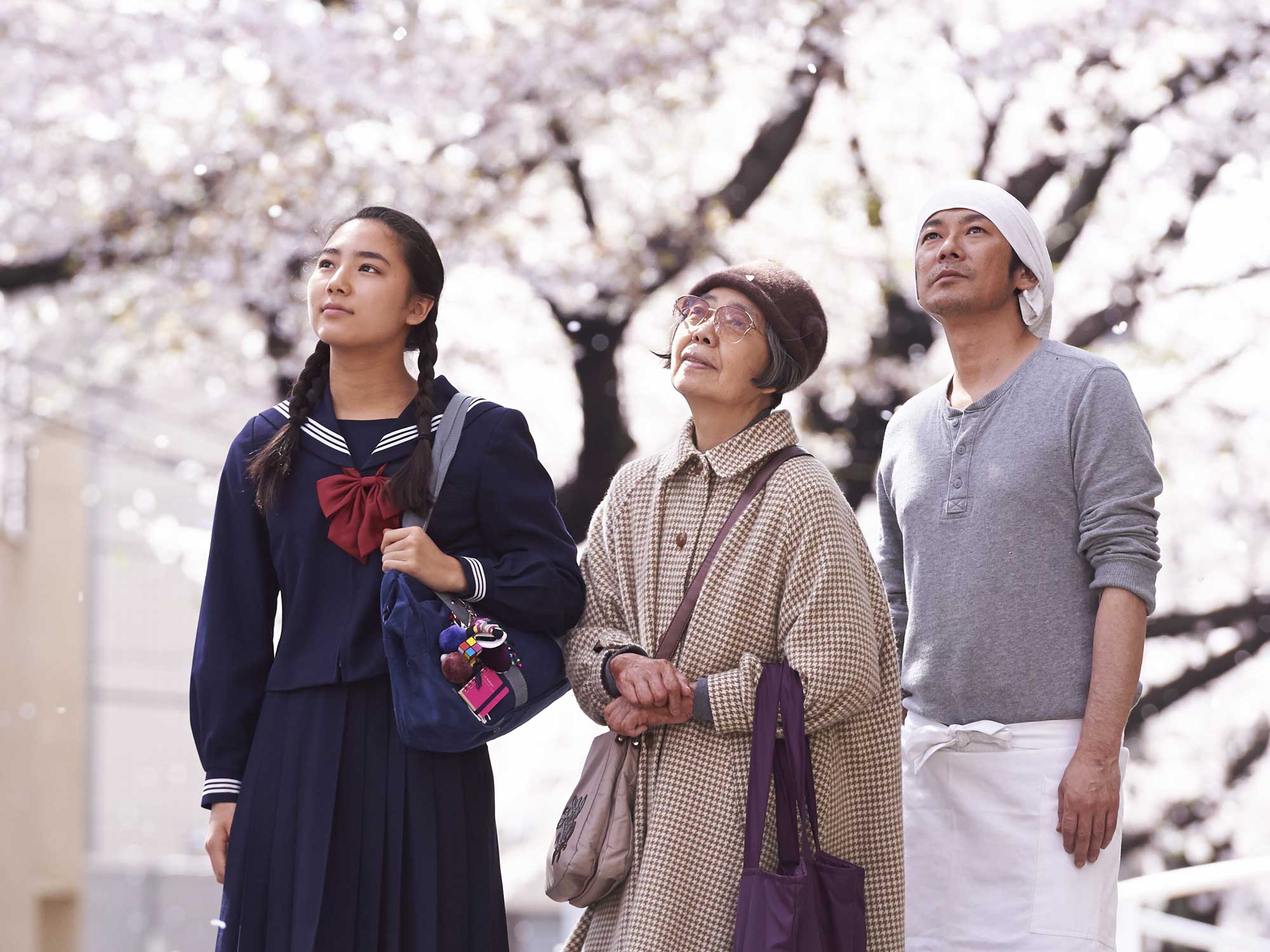 Image is from the film 'Sweet Bean' (2015). A school girl, an old woman, and a man all stand together and watch cherry blossoms fall from their branches.
